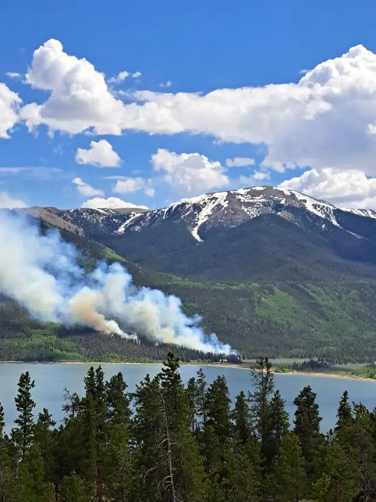 Scenic landscape with lake, forest, mountains, and a wildfire.