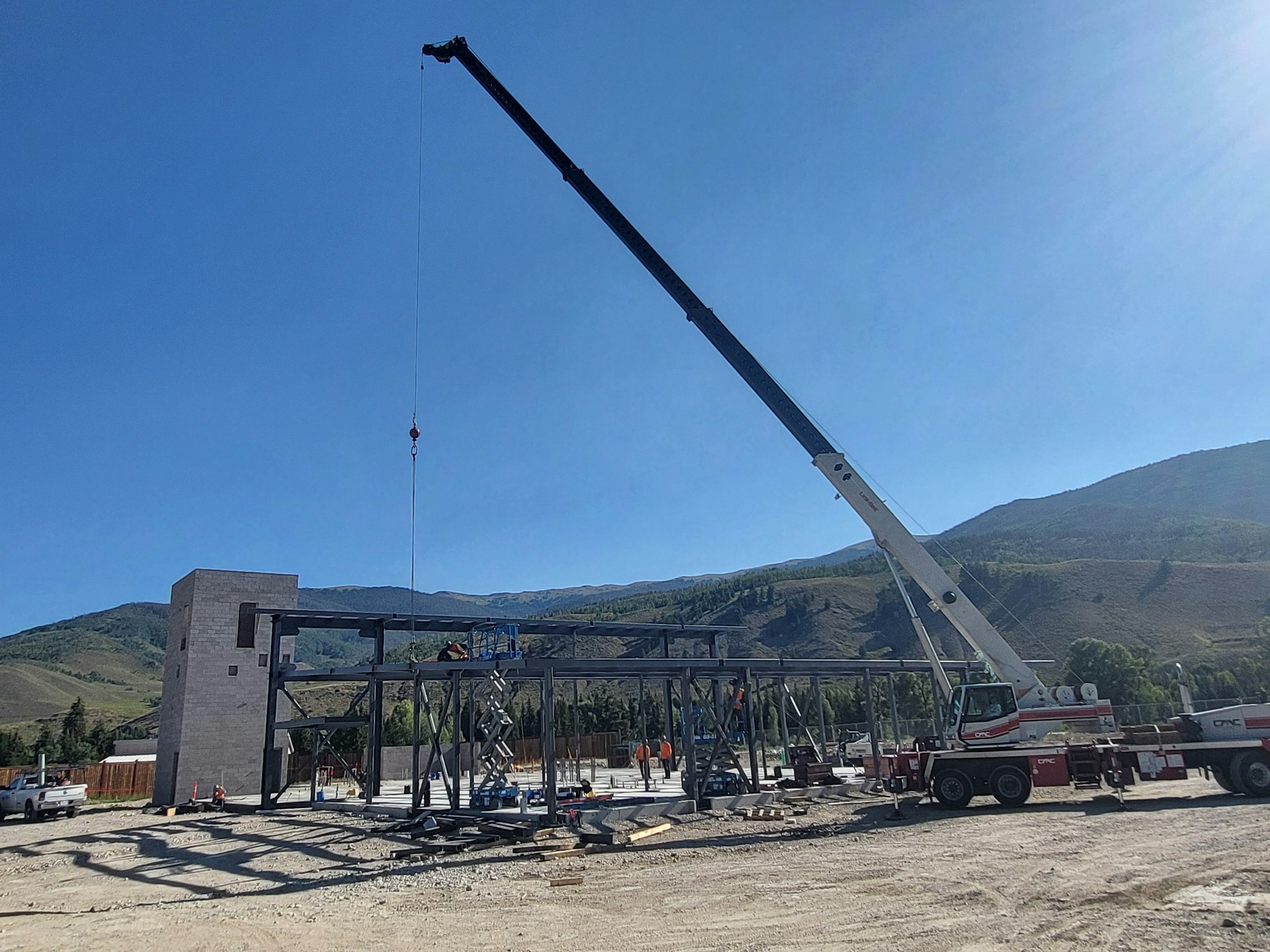 A construction site with a crane lifting steel beams, sunny sky, workers, and mountains in the background.