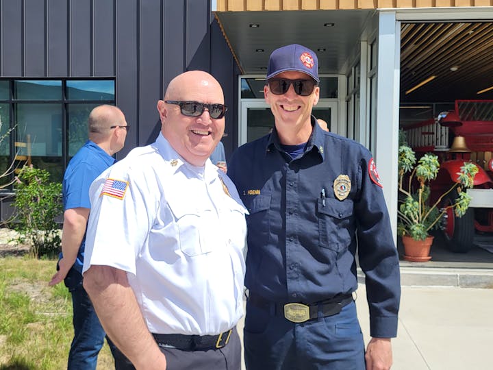 Two uniformed firefighters smiling outside a building with a fire truck inside.