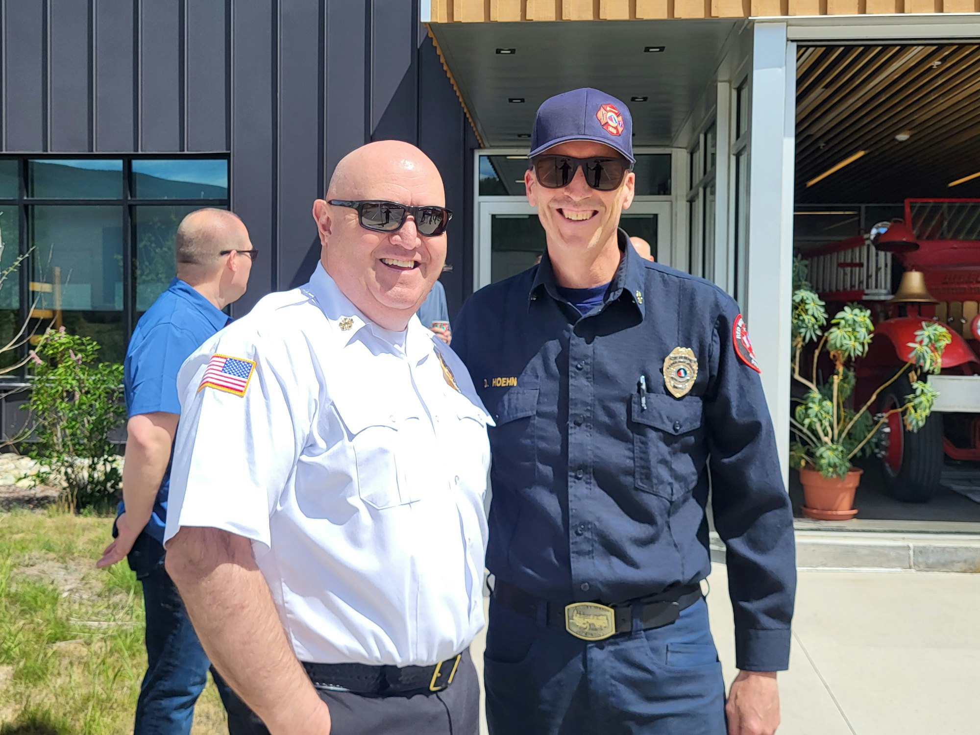 Two uniformed firefighters smiling outside a building with a fire truck inside.