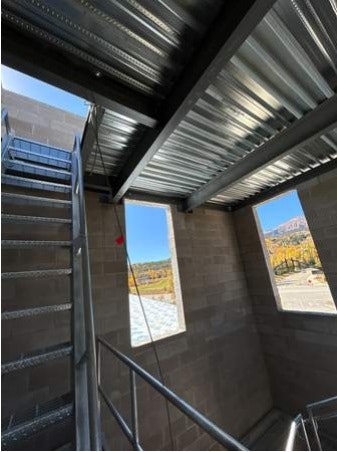 Interior view of a stairwell with metal steps and corrugated ceiling, looking out to a bright day through a window.