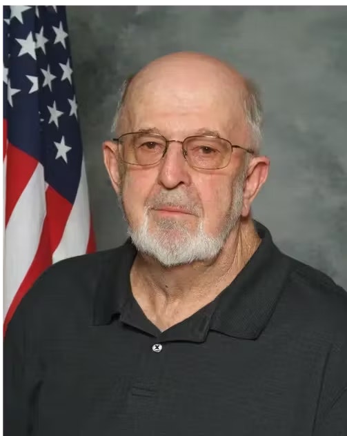 An older man with glasses and a beard in a collared shirt stands in front of an American flag.
