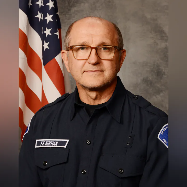 A man in a fire department uniform poses in front of an American flag. He has glasses and a serious expression.