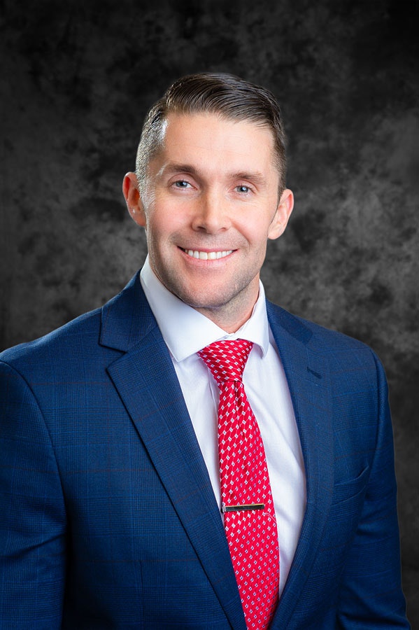 Man in a blue suit with a red tie, smiling against a dark textured background.