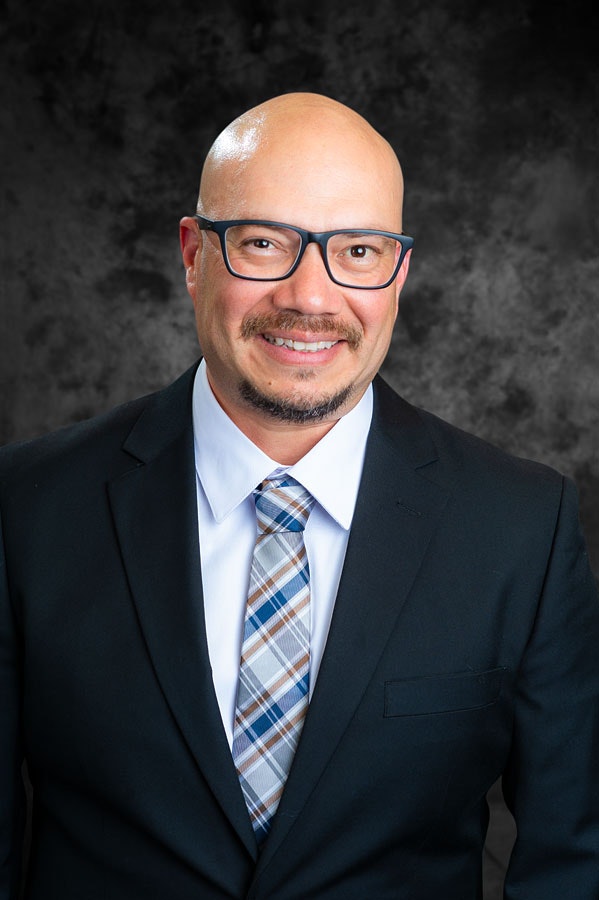 A person wearing glasses, a suit, and a patterned tie, smiling against a dark gray background.