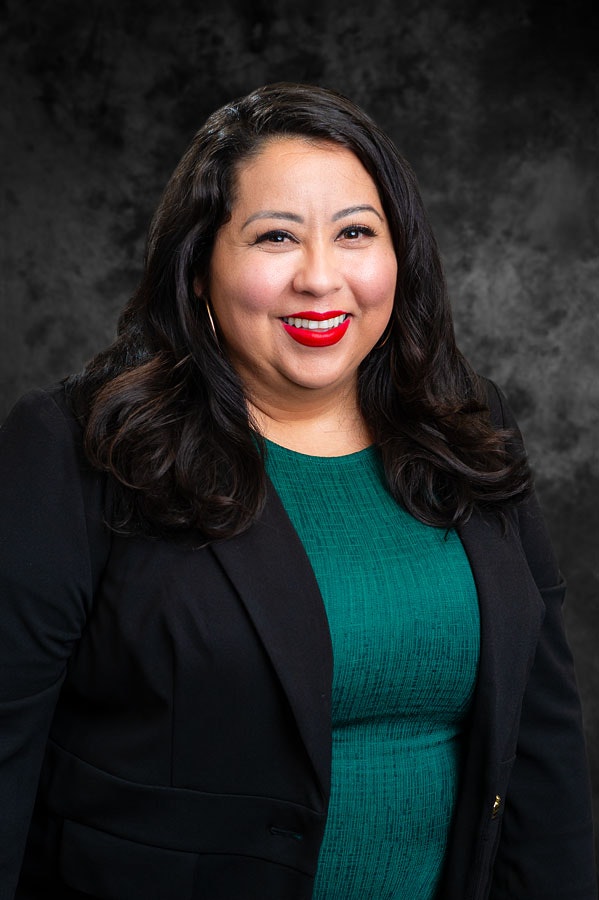 A person in a green dress and black blazer smiles against a dark gray backdrop.