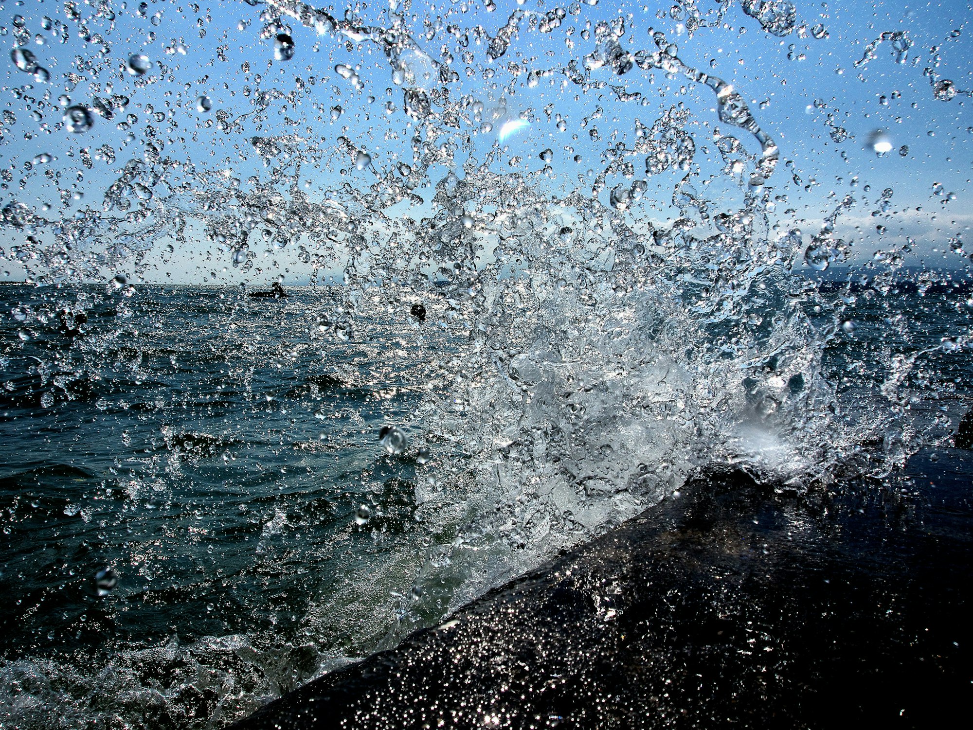 Waves crashing against rocks, water droplets suspended in the air, with a clear blue sky in the background.