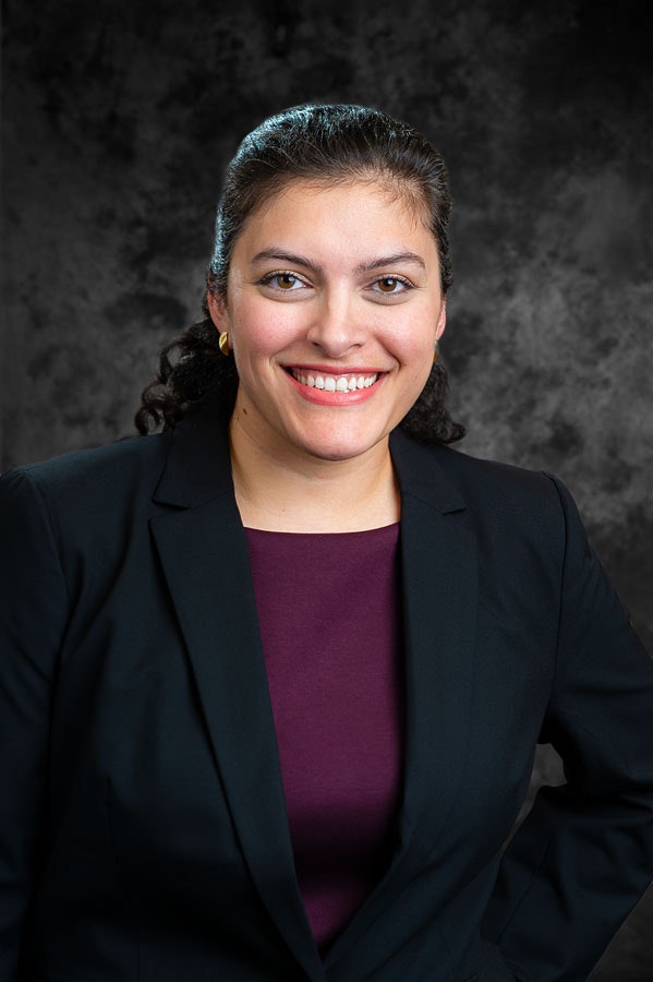 A person in formal attire, smiling, with a dark gray textured background.