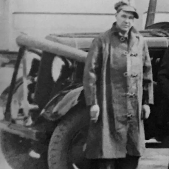 A man wearing a long coat and hat stands beside a vintage car, likely from an earlier era.