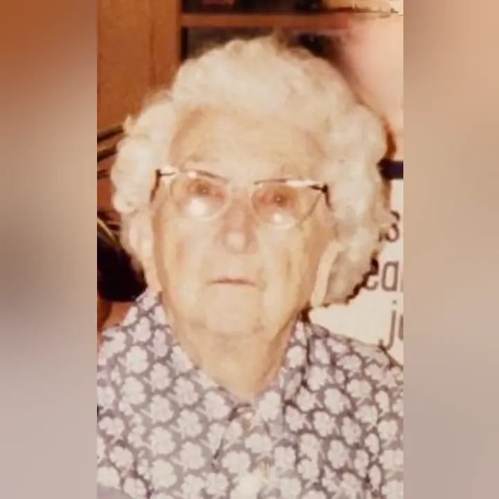 An elderly woman with curly white hair and glasses is looking directly at the camera, wearing a floral-patterned shirt.