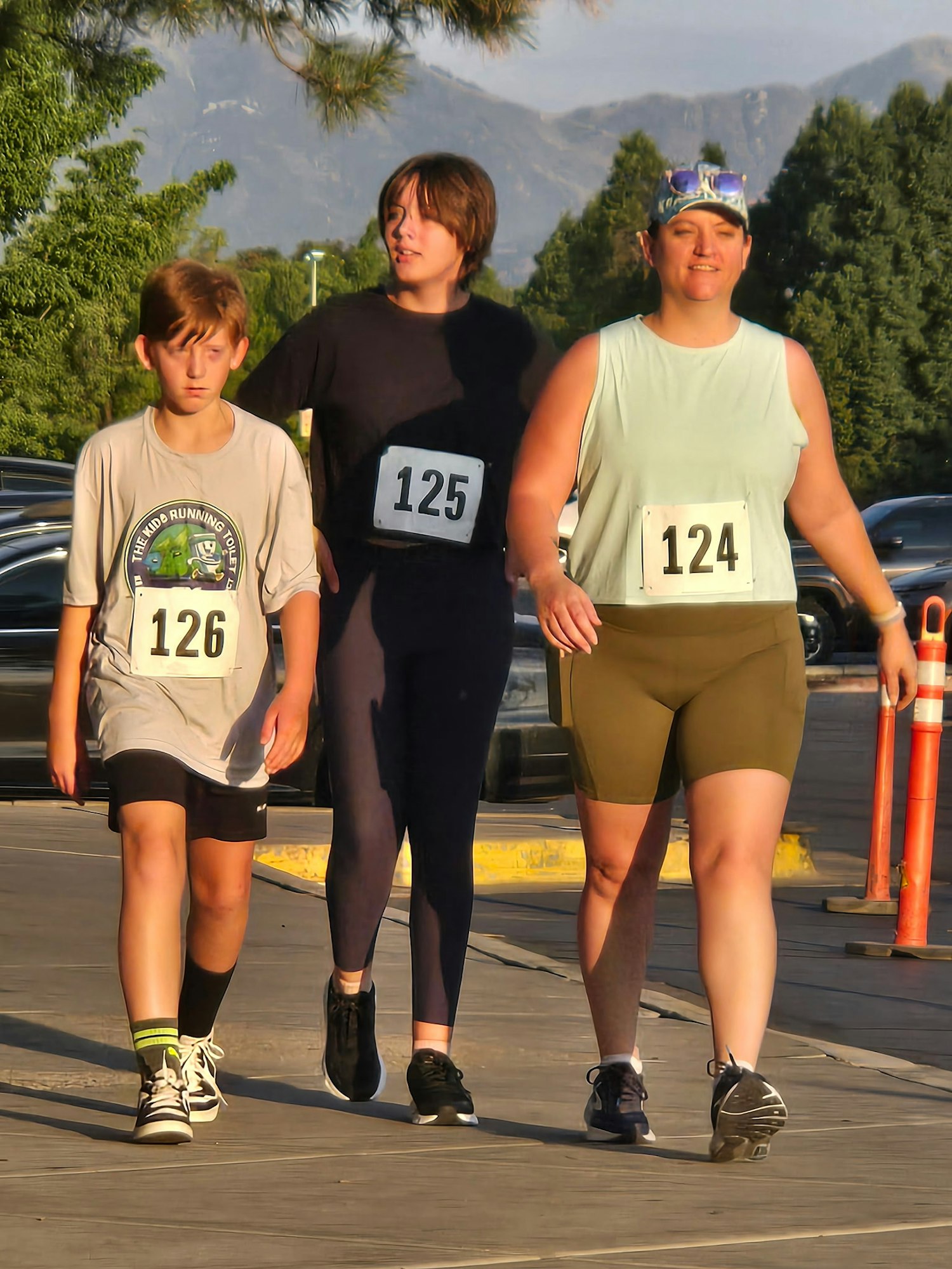 Three people walk together outdoors, wearing numbered race bibs. The setting has trees and mountains in the background.