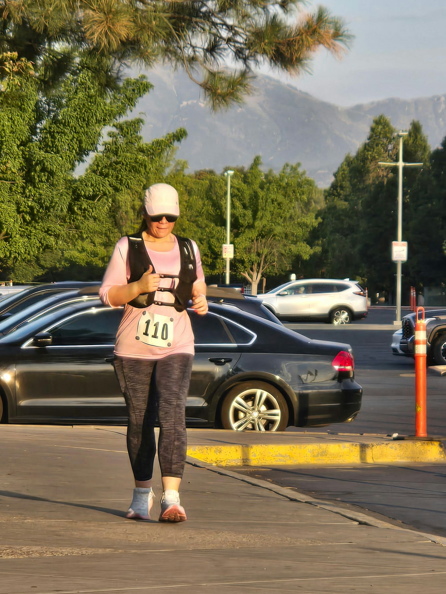 A woman in athletic wear with a race number is walking along a parking lot with cars and trees in the background.