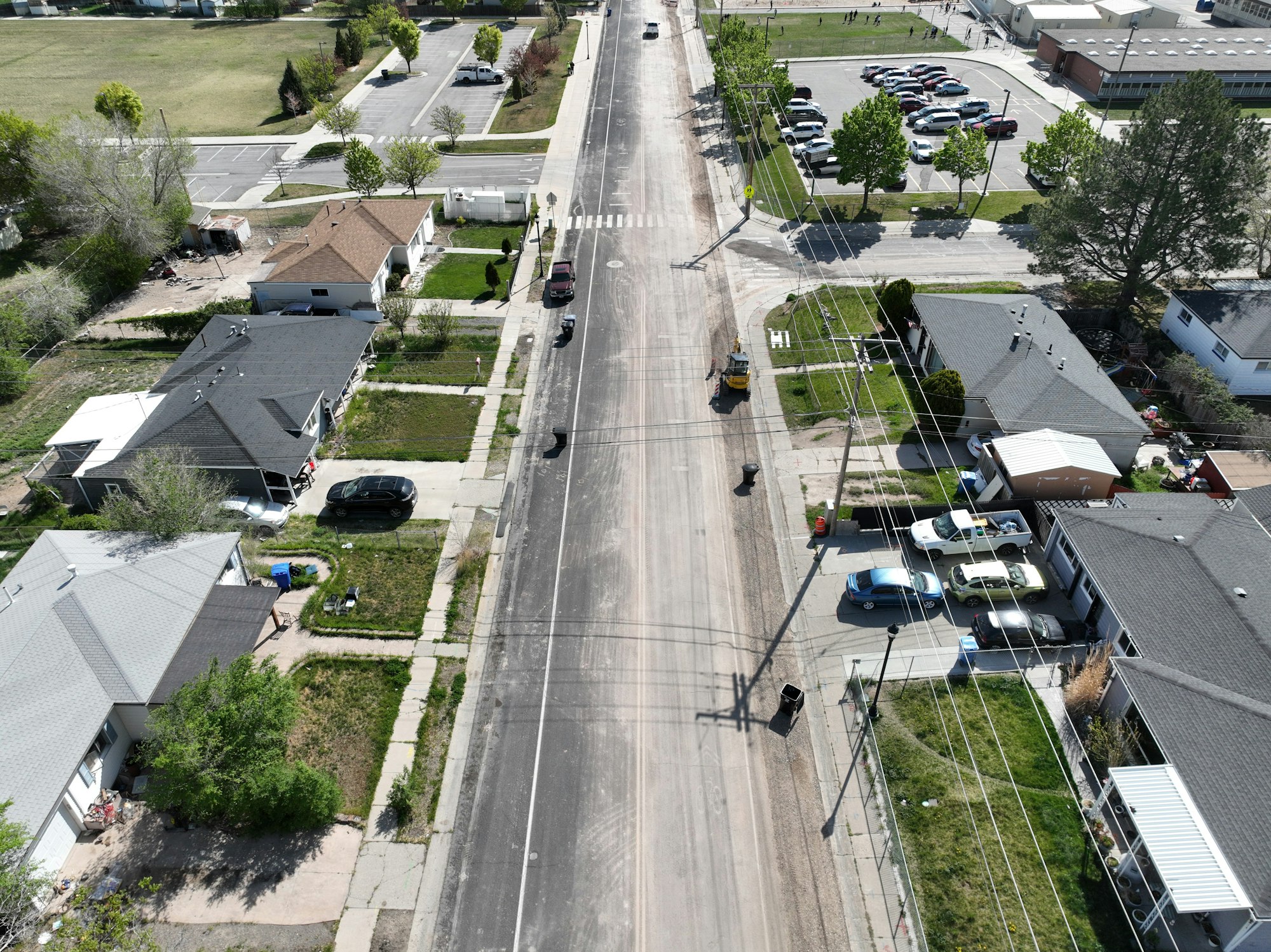 An aerial view of residential homes, a road, and a parking lot with multiple cars, showing a mix of greenery and urban setting.