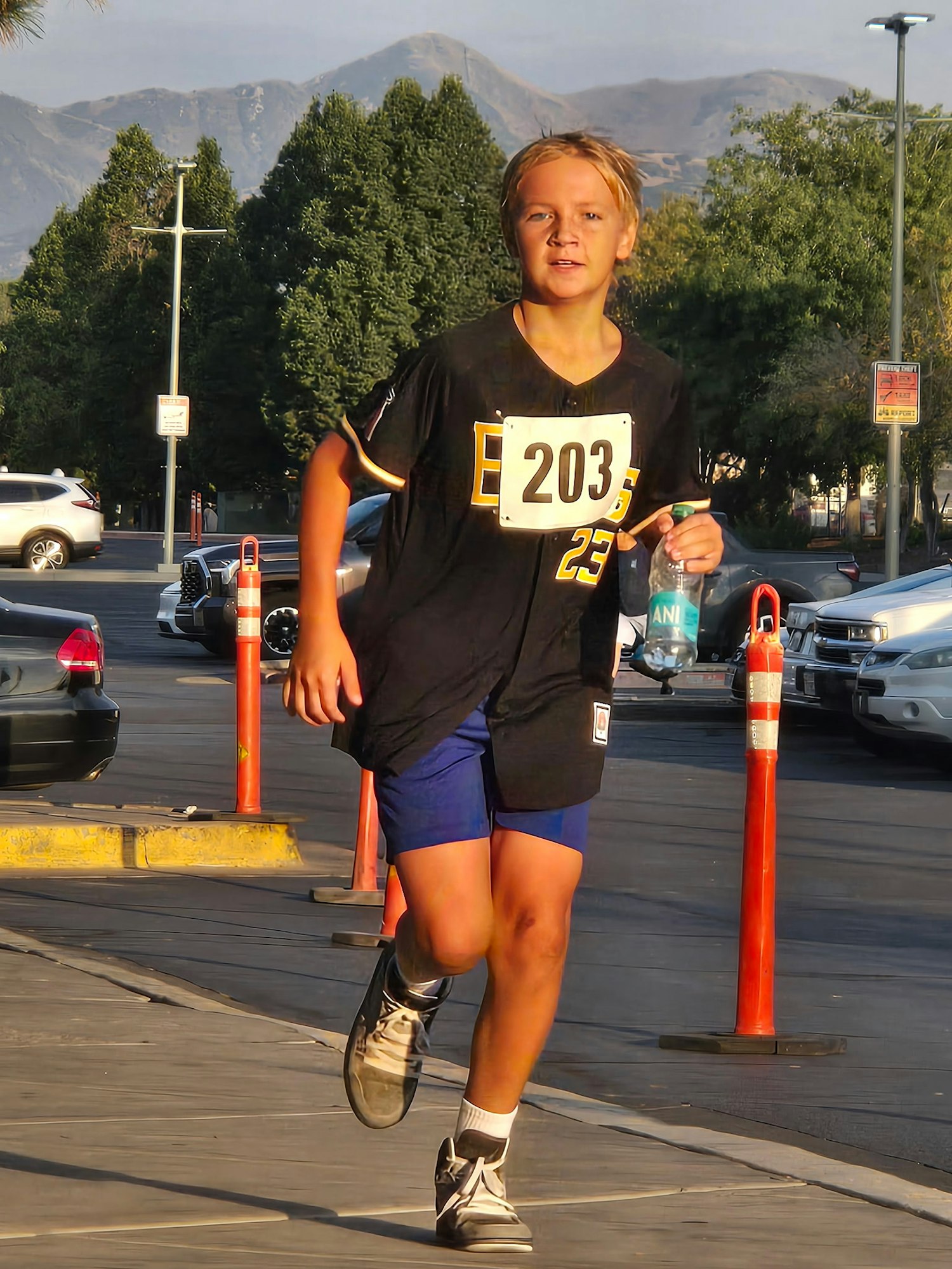 A young runner wearing a jersey with the number 203 jogs with a water bottle in a parking area, mountains in the background.