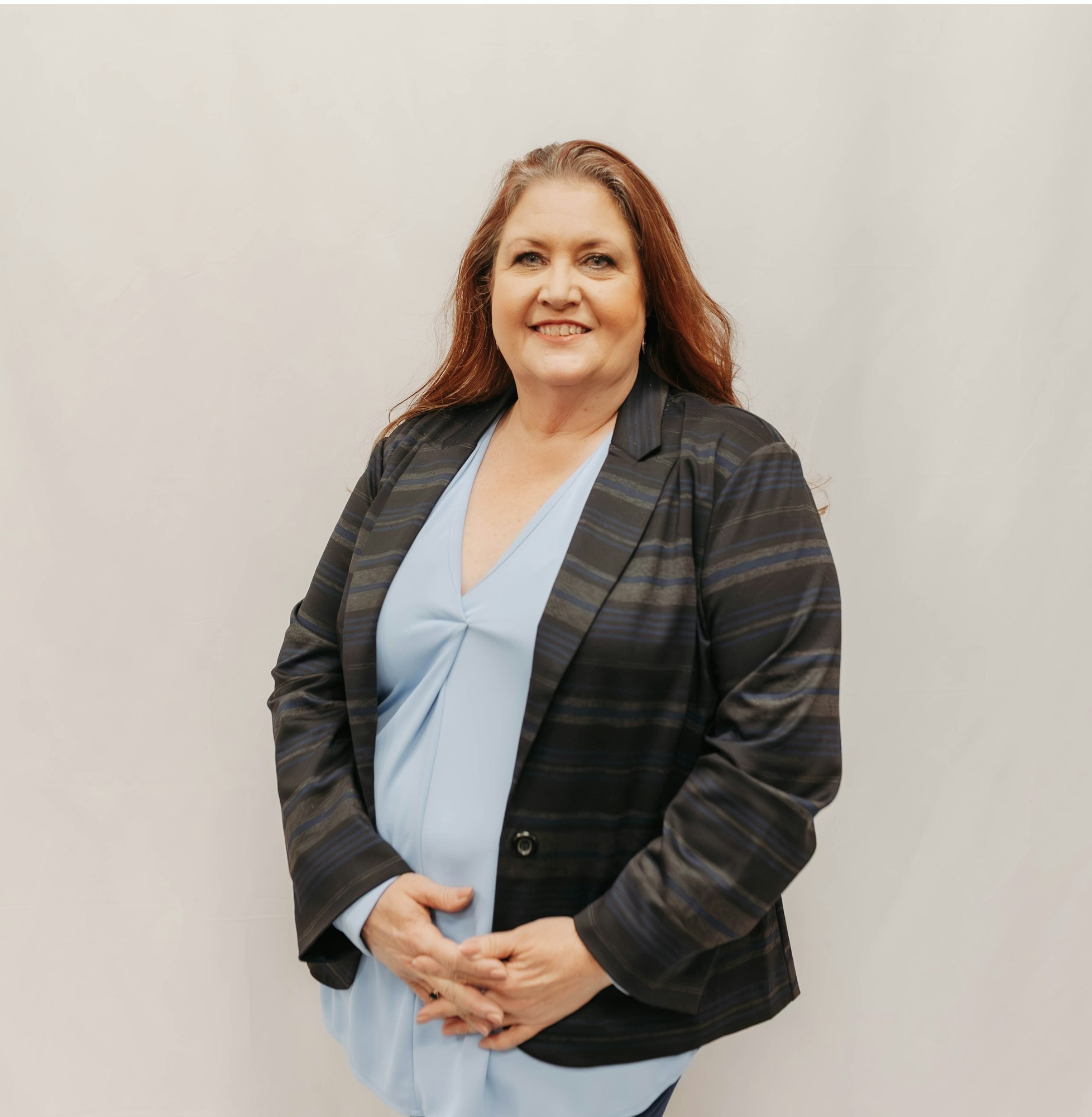 The image shows a woman with long hair, wearing a black striped blazer over a light blue top, posing against a neutral background.