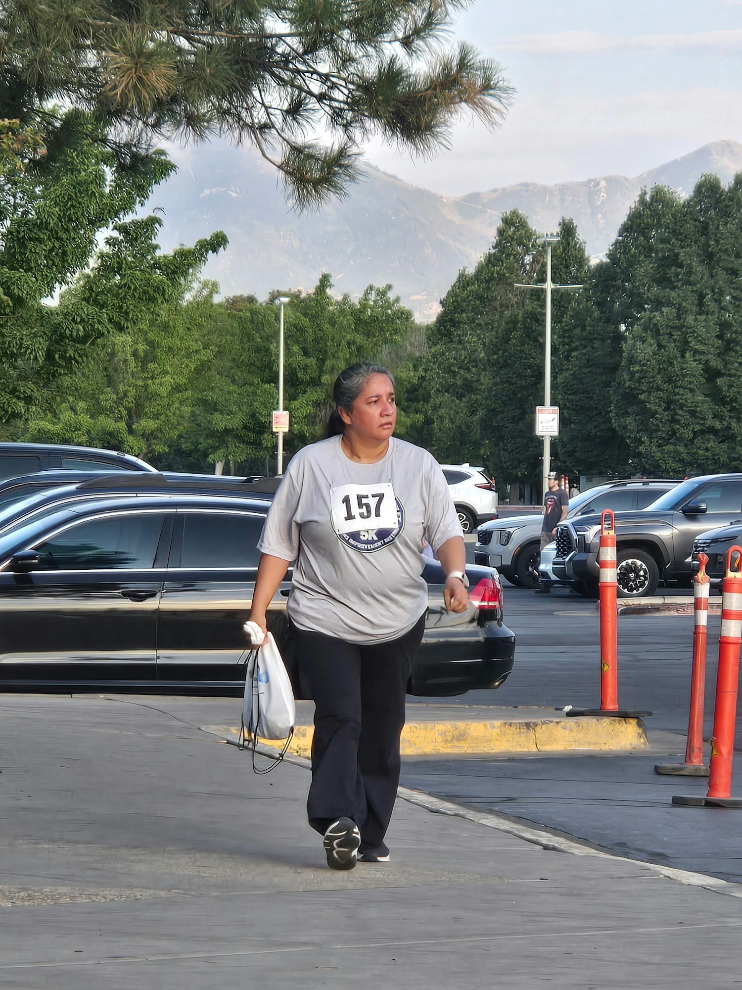A woman in a gray shirt with a number walks casually near parked cars and trees, with mountains in the background.