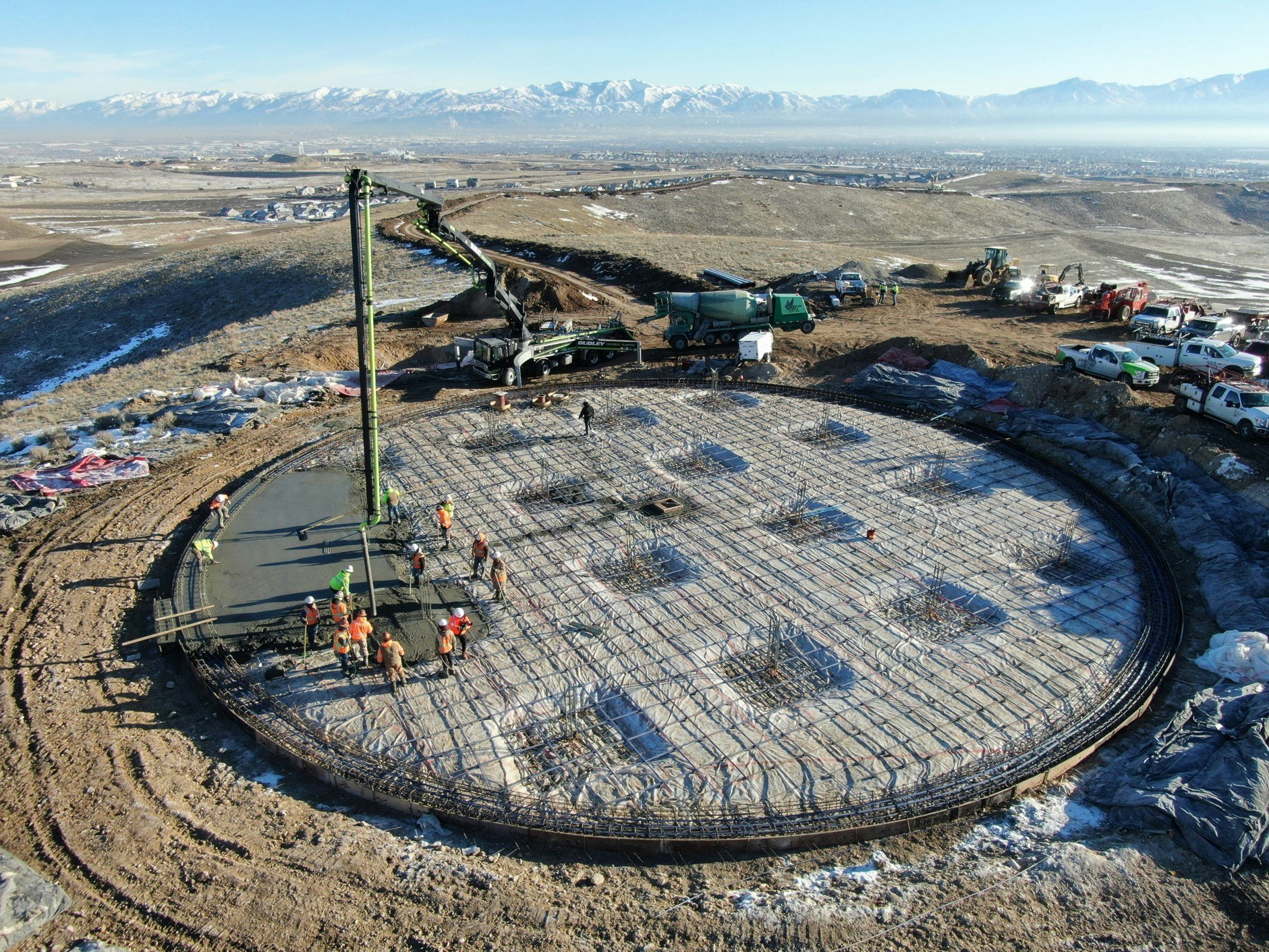Construction site with workers pouring concrete on a circular base, surrounded by machinery and a mountainous backdrop.