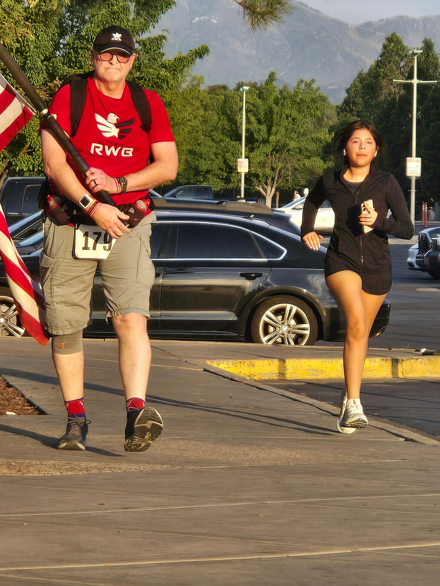 A man with a flag and a number on his shirt walks while a woman jogs past him in a sunny outdoor setting.