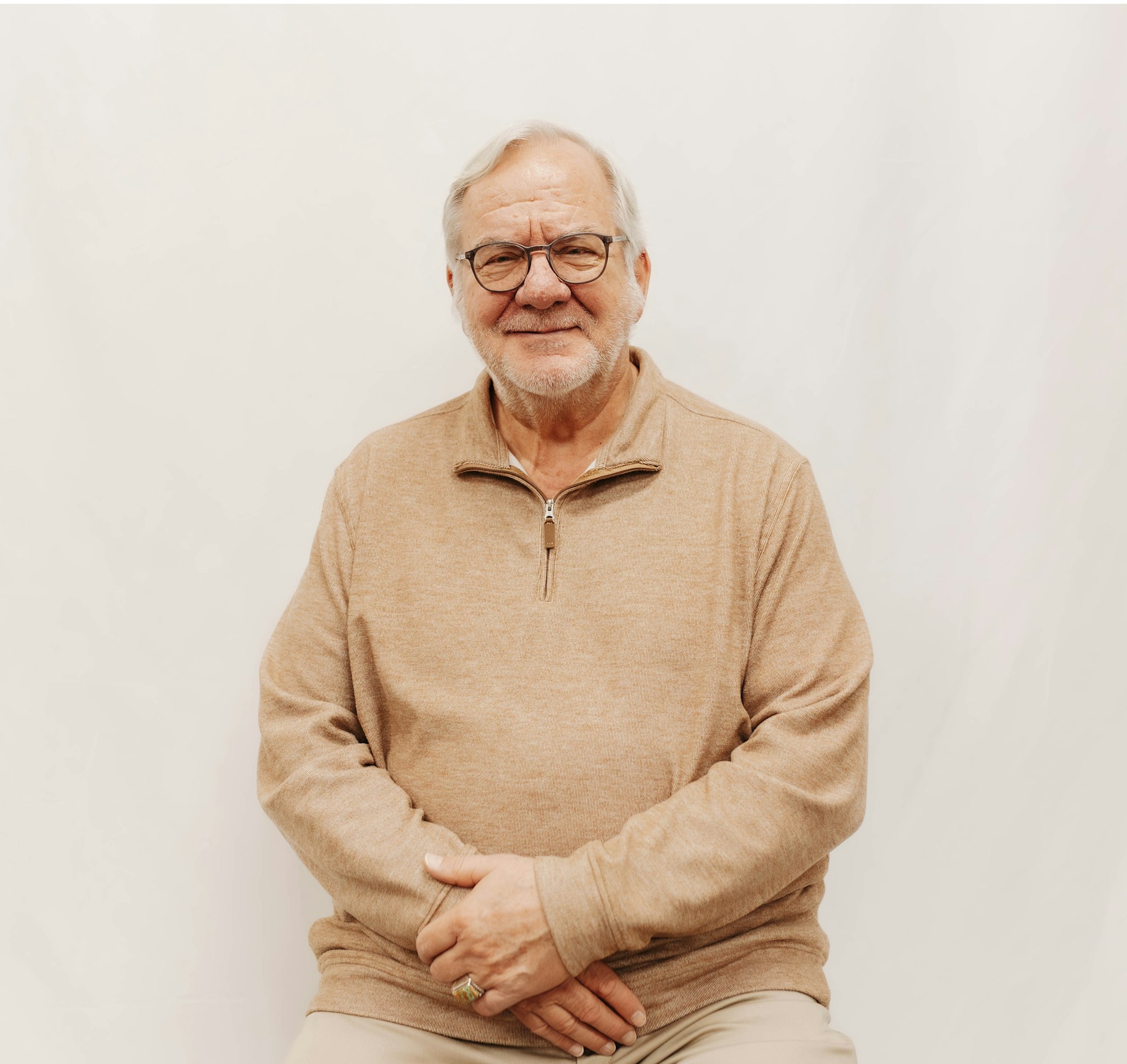 A smiling older man with glasses, wearing a tan sweater, seated against a plain light-colored background.
