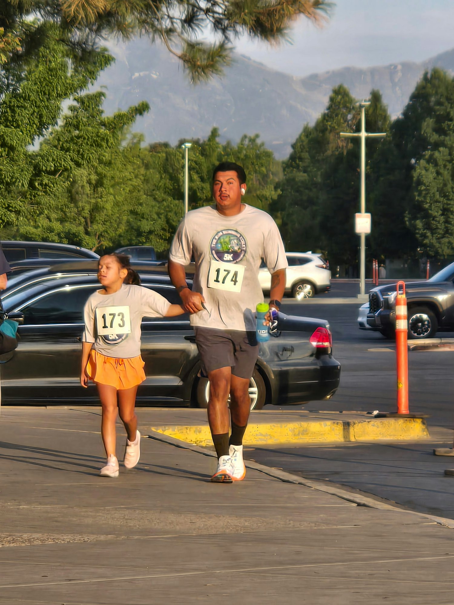 A man and a girl run together, each wearing a race bib, in a sunny outdoor setting with trees and cars around them.