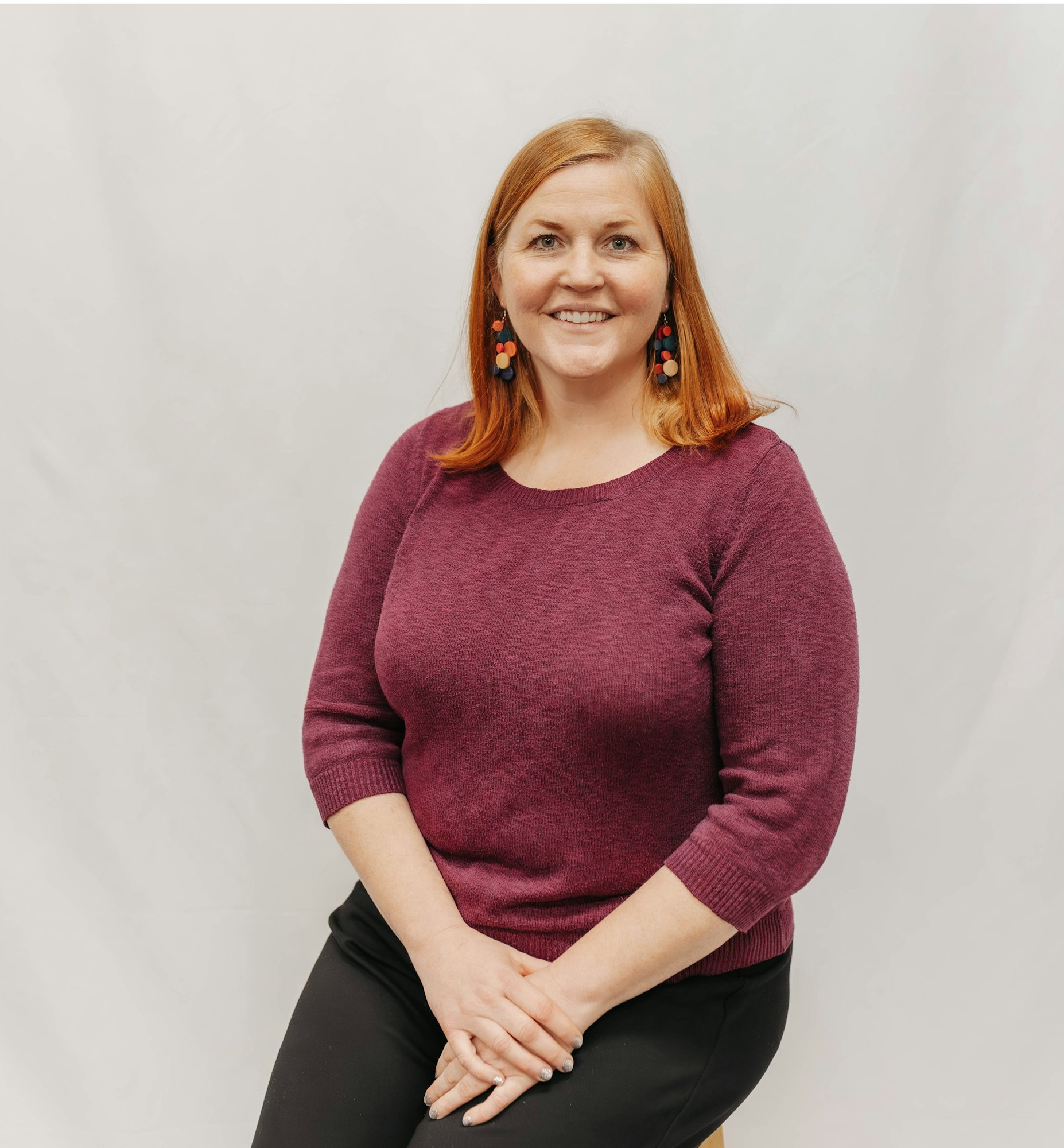 A smiling woman with red hair sits against a light background, wearing a purple sweater and colorful earrings.
