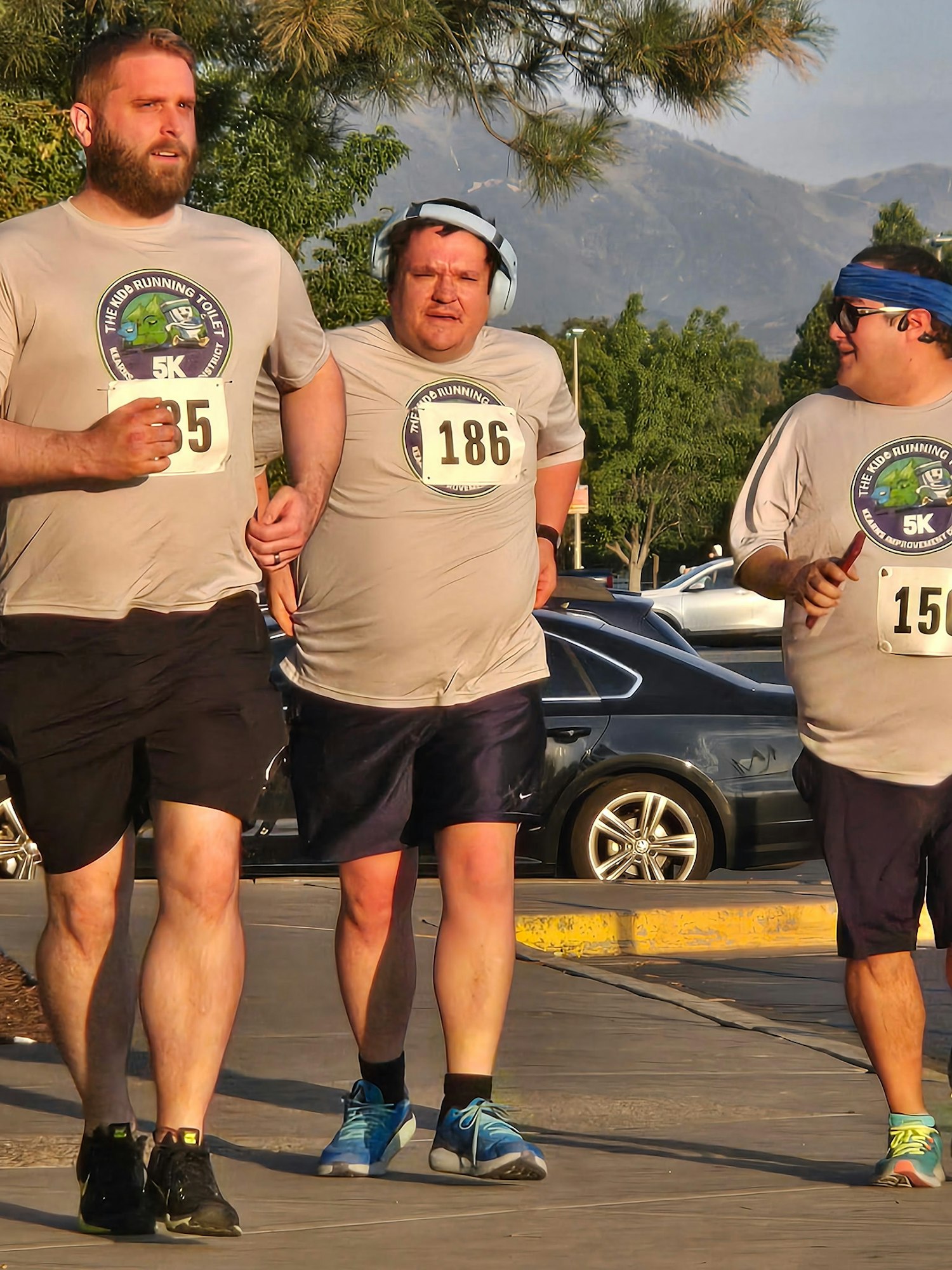 Three men, wearing matching shirts with numbers, are jogging together in a sunny outdoor setting, surrounded by trees and cars.