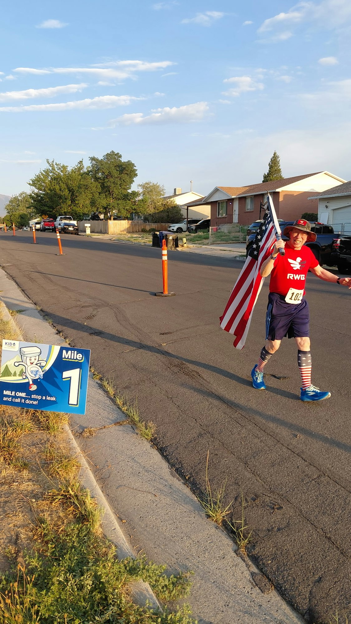A runner wearing a flag-themed outfit carries an American flag while passing a "Mile 1" sign on a residential street.