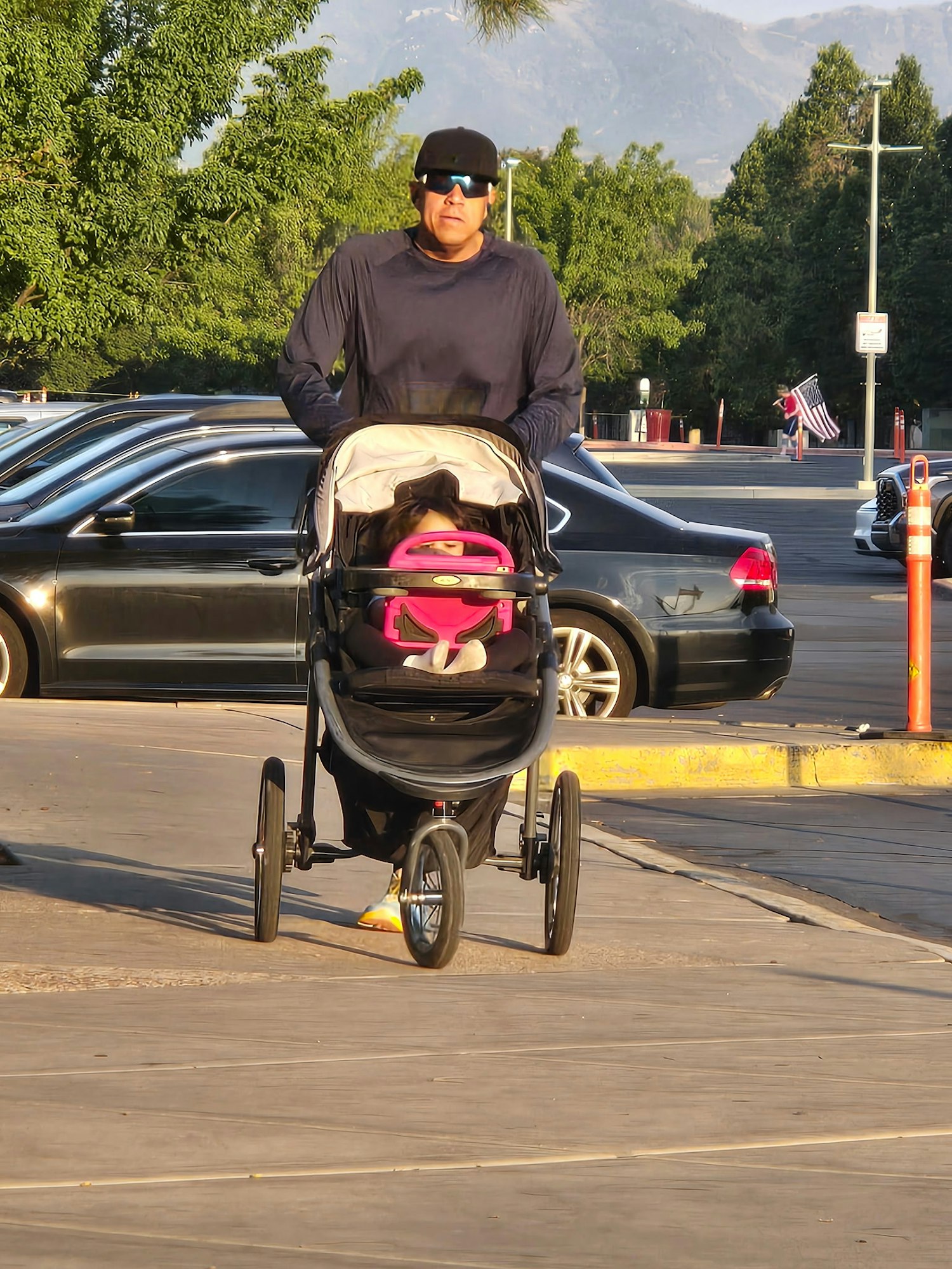 A man pushes a stroller with a child inside, walking near parked cars and trees in a suburban setting.