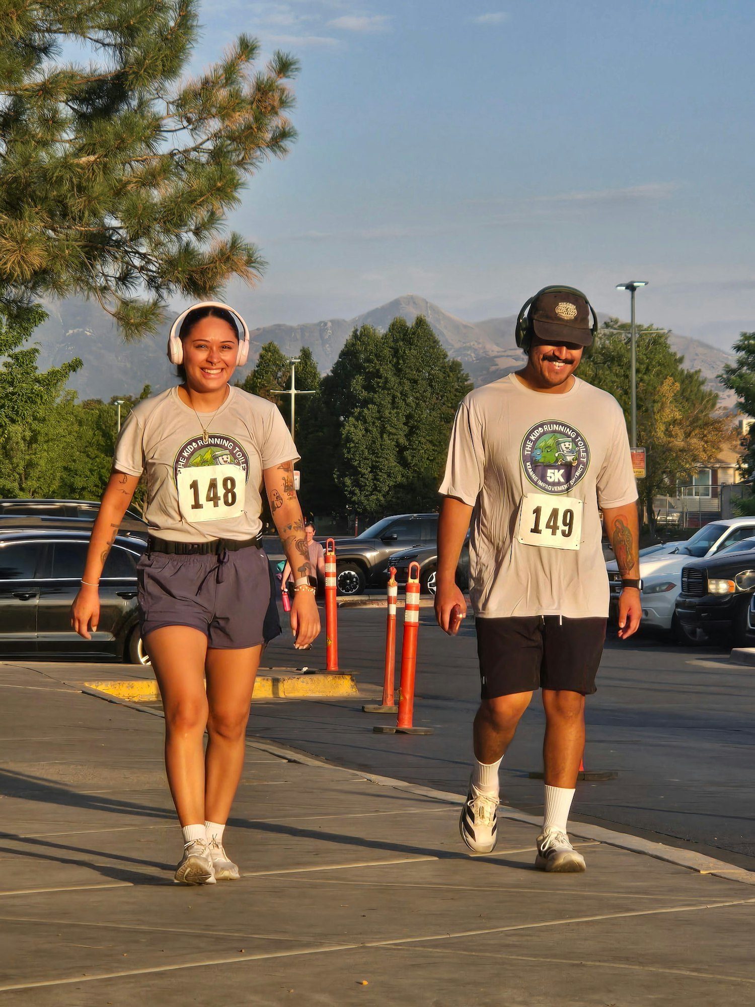 Two people are walking in sports attire, wearing headphones and numbered bibs, amidst a scenic backdrop with trees and mountains.