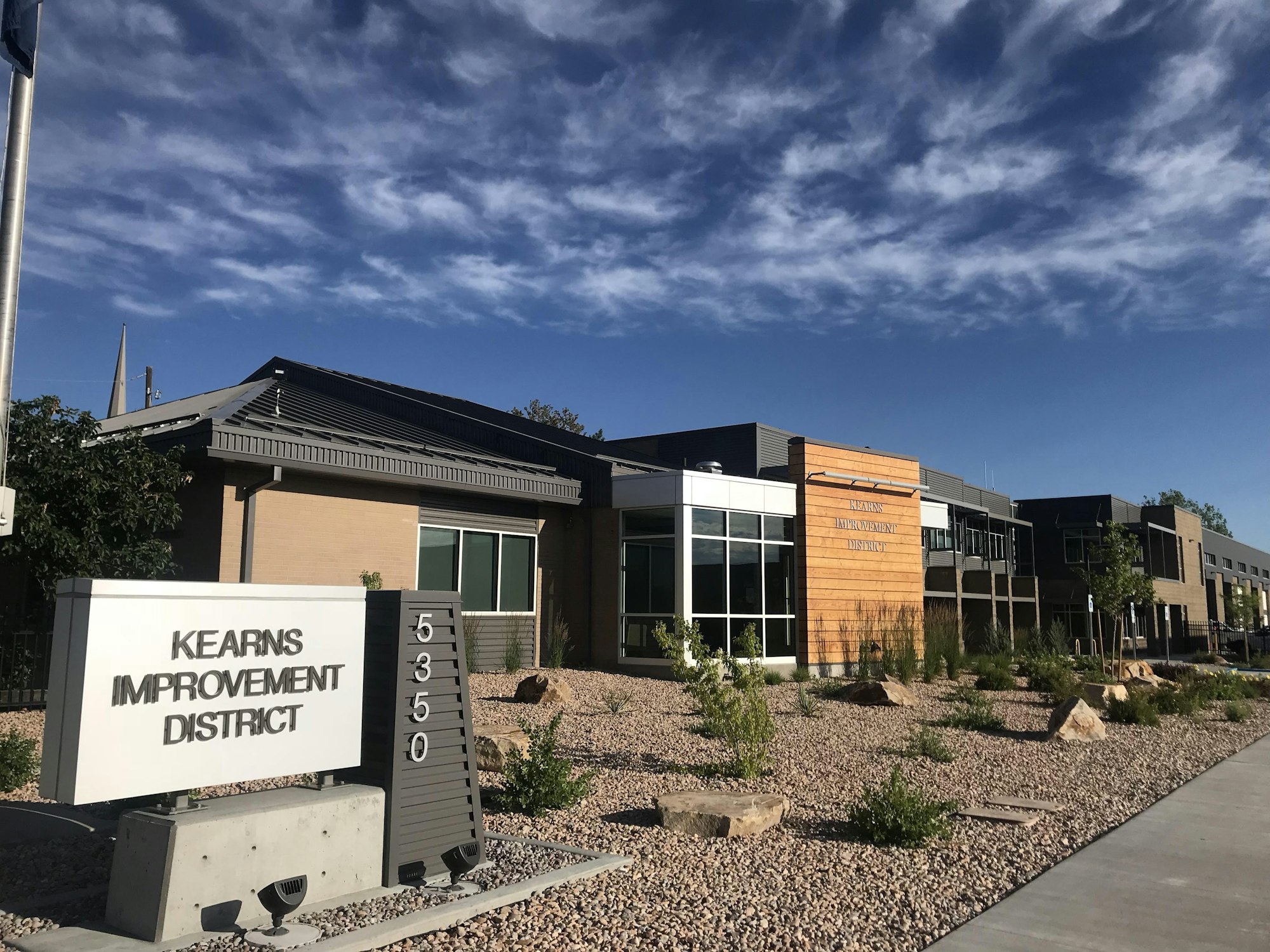 Modern building with sign "Kearns Improvement District," landscaping, blue sky with wispy clouds.