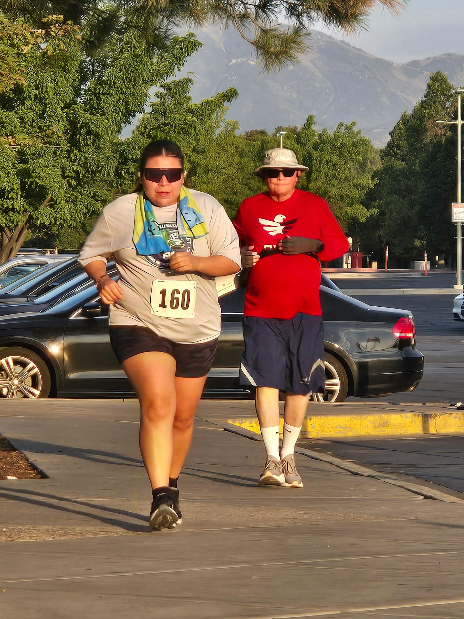 Two runners are jogging on a path, one in athletic gear with a race number, while a man in a red shirt follows. Trees and cars are in the background.