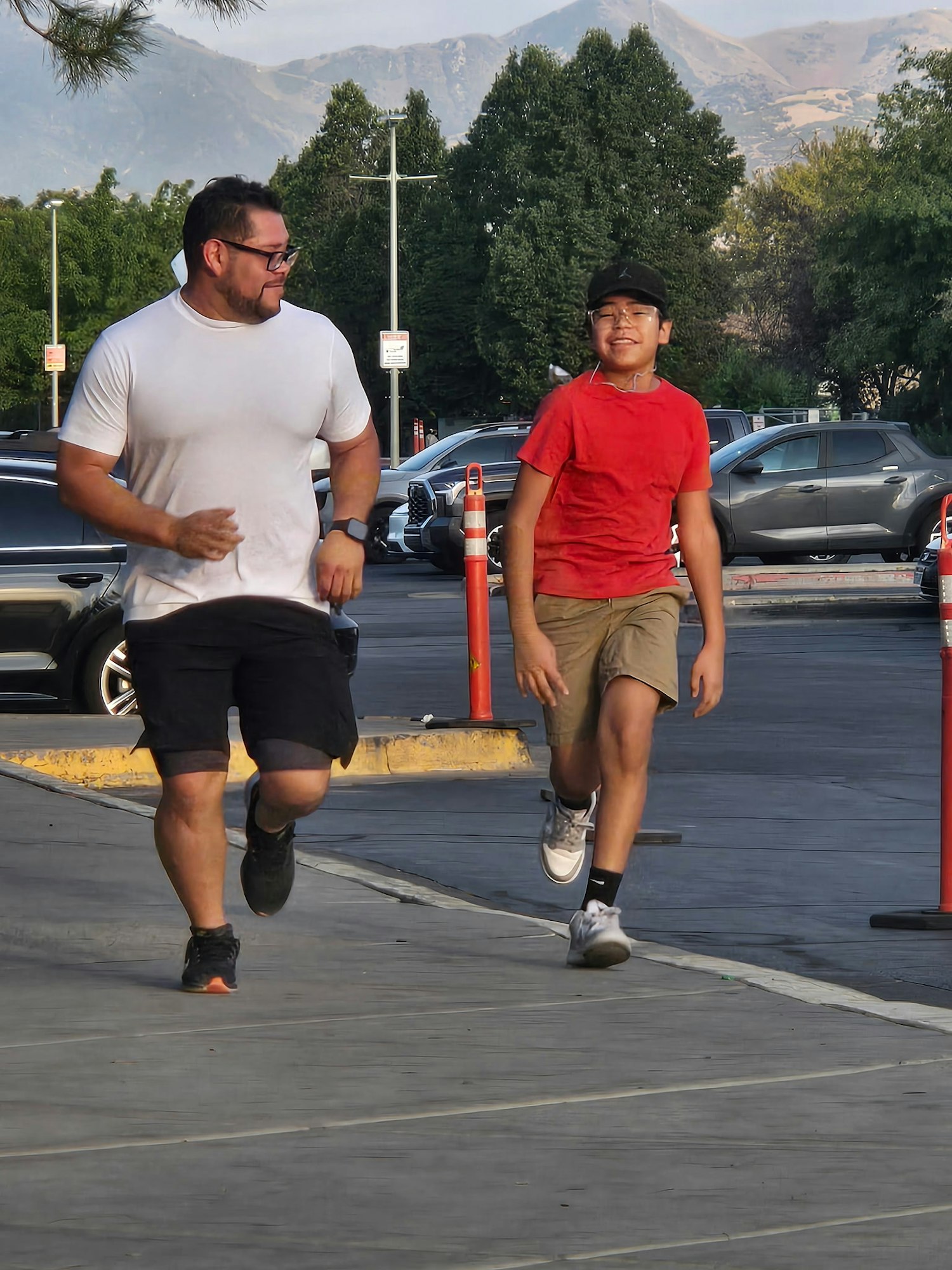 A man and a boy are jogging together on a sidewalk, smiling, surrounded by trees and parked cars in the background.