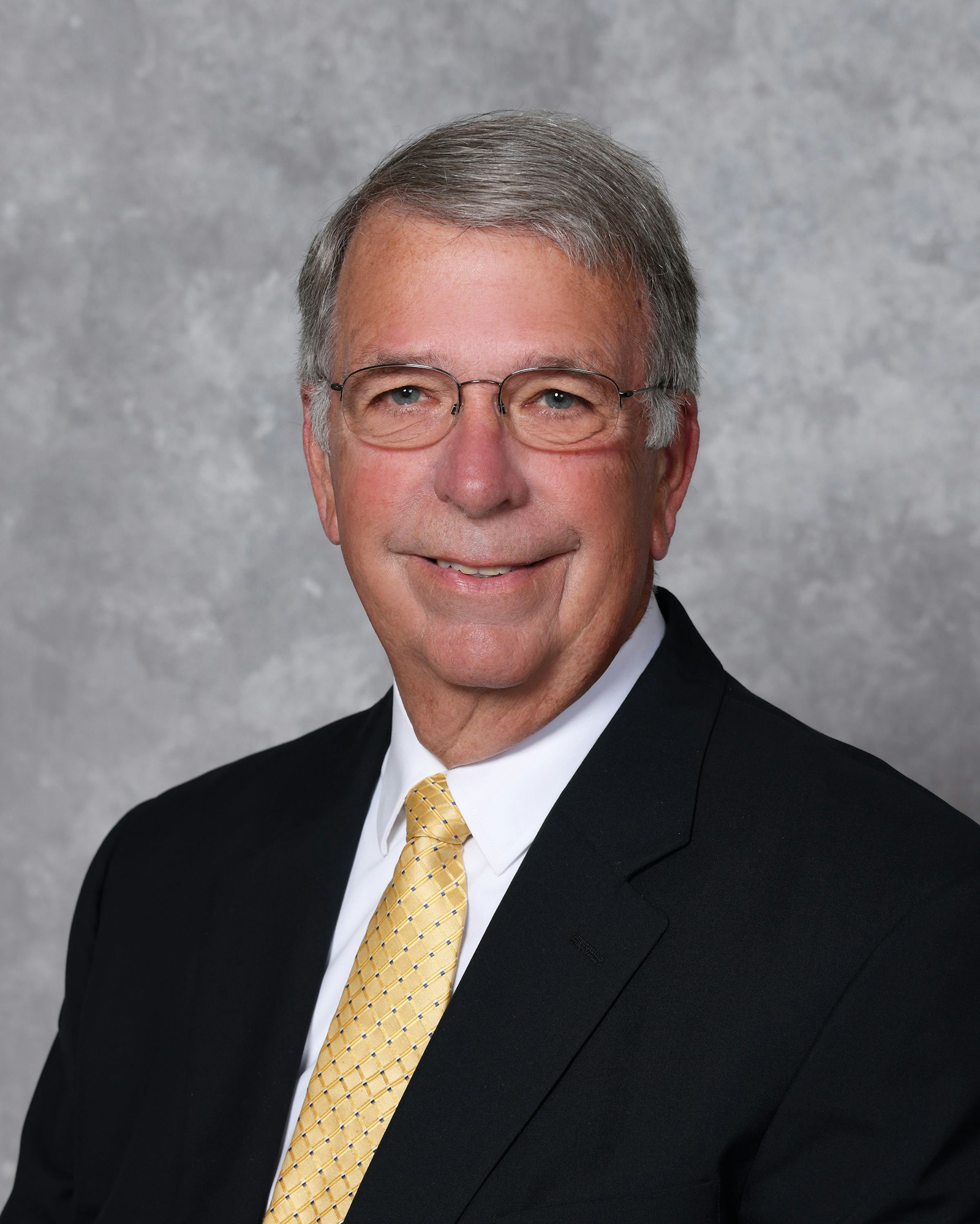Portrait of a smiling man in a suit and patterned tie against a gray background.