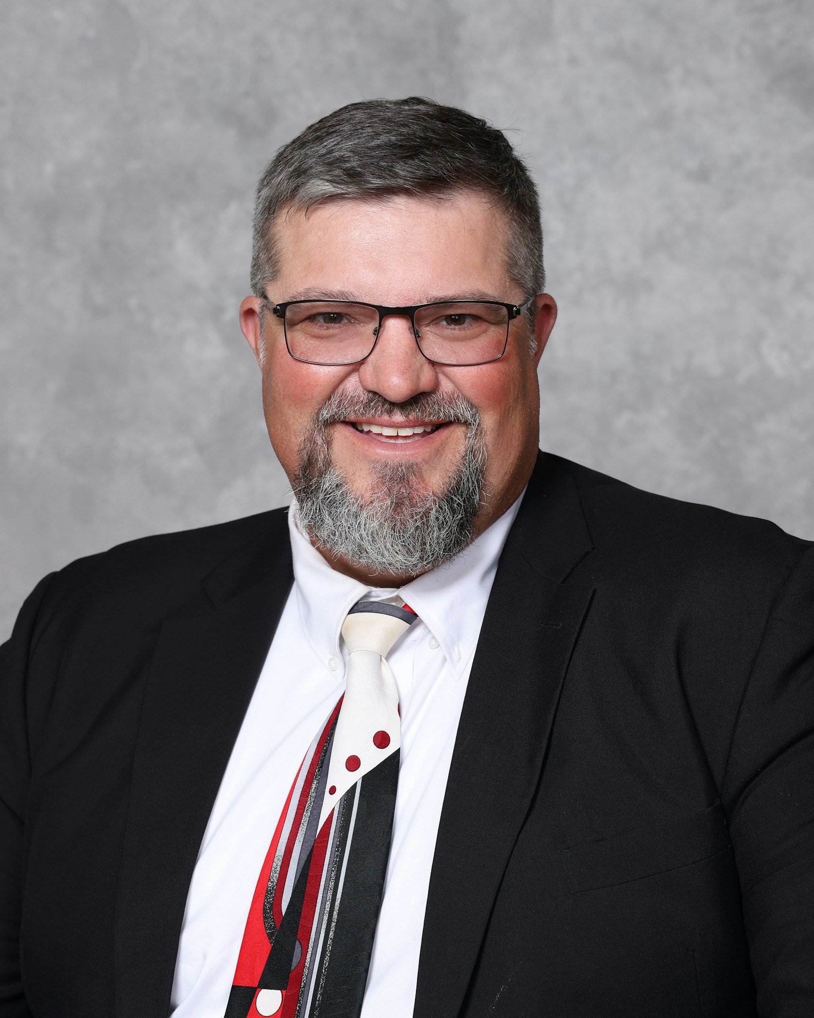Smiling man with glasses, beard, suit, and artistic tie.