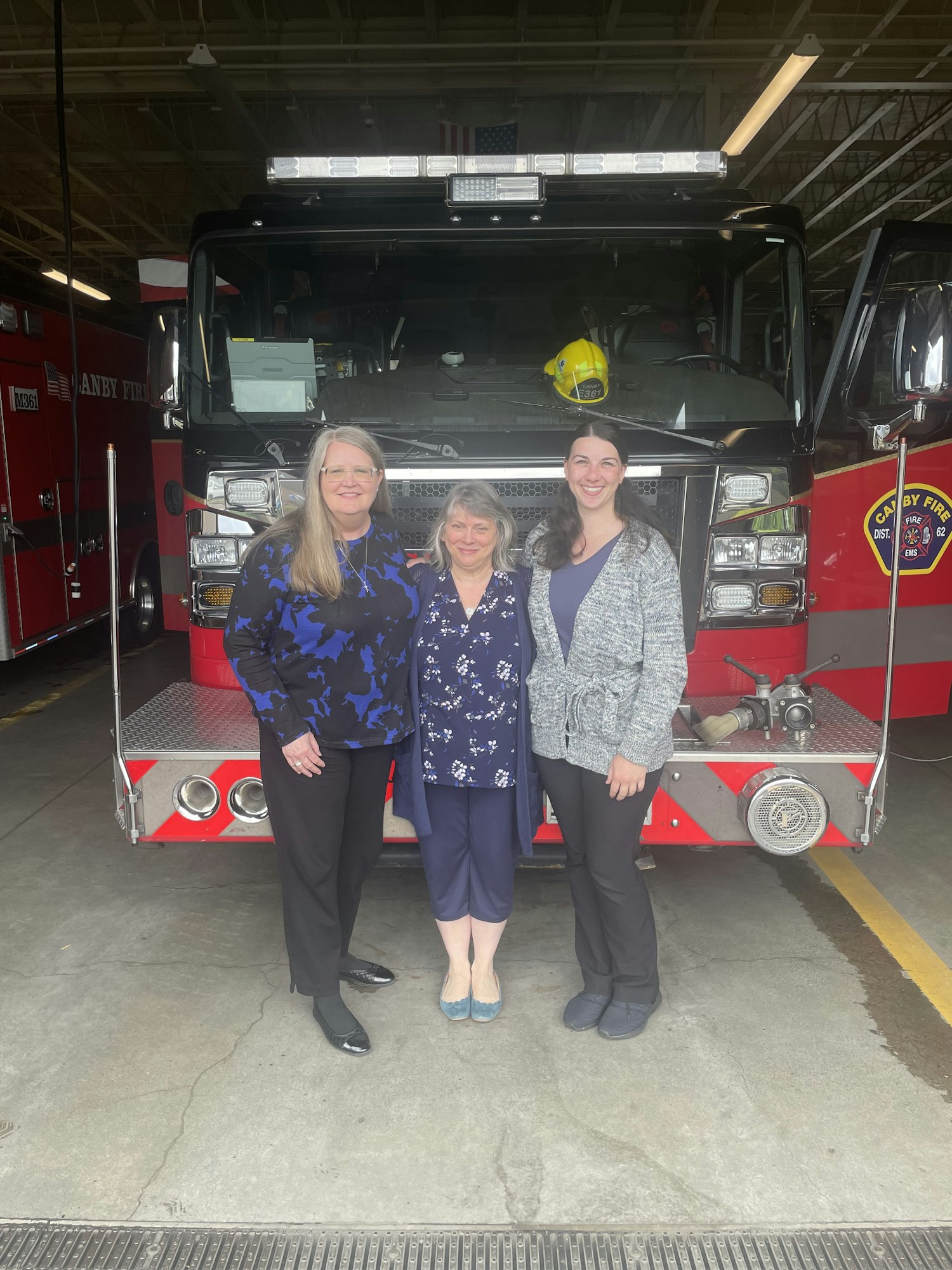 Three people standing in front of a fire truck inside a fire station.
