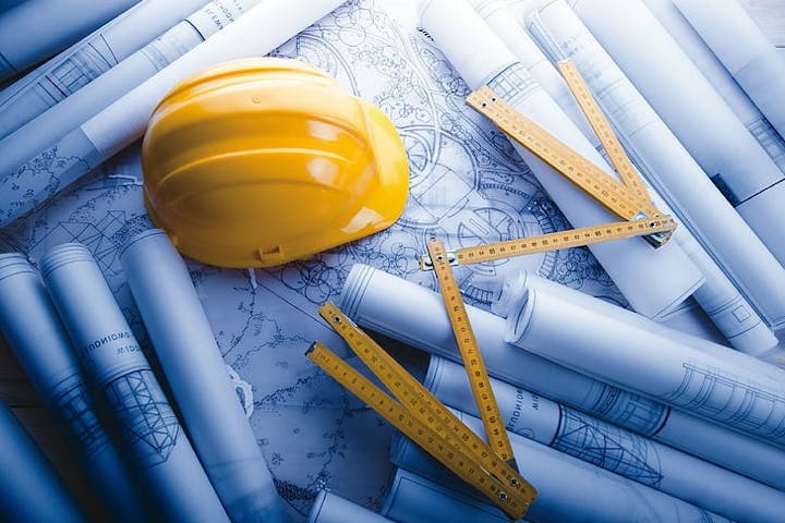 A yellow hard hat, rolled engineering blueprints, and a folding ruler on a table.