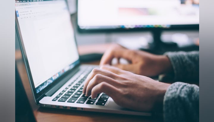 A person typing on a laptop, with another screen visible in the background, likely working on a document or project.