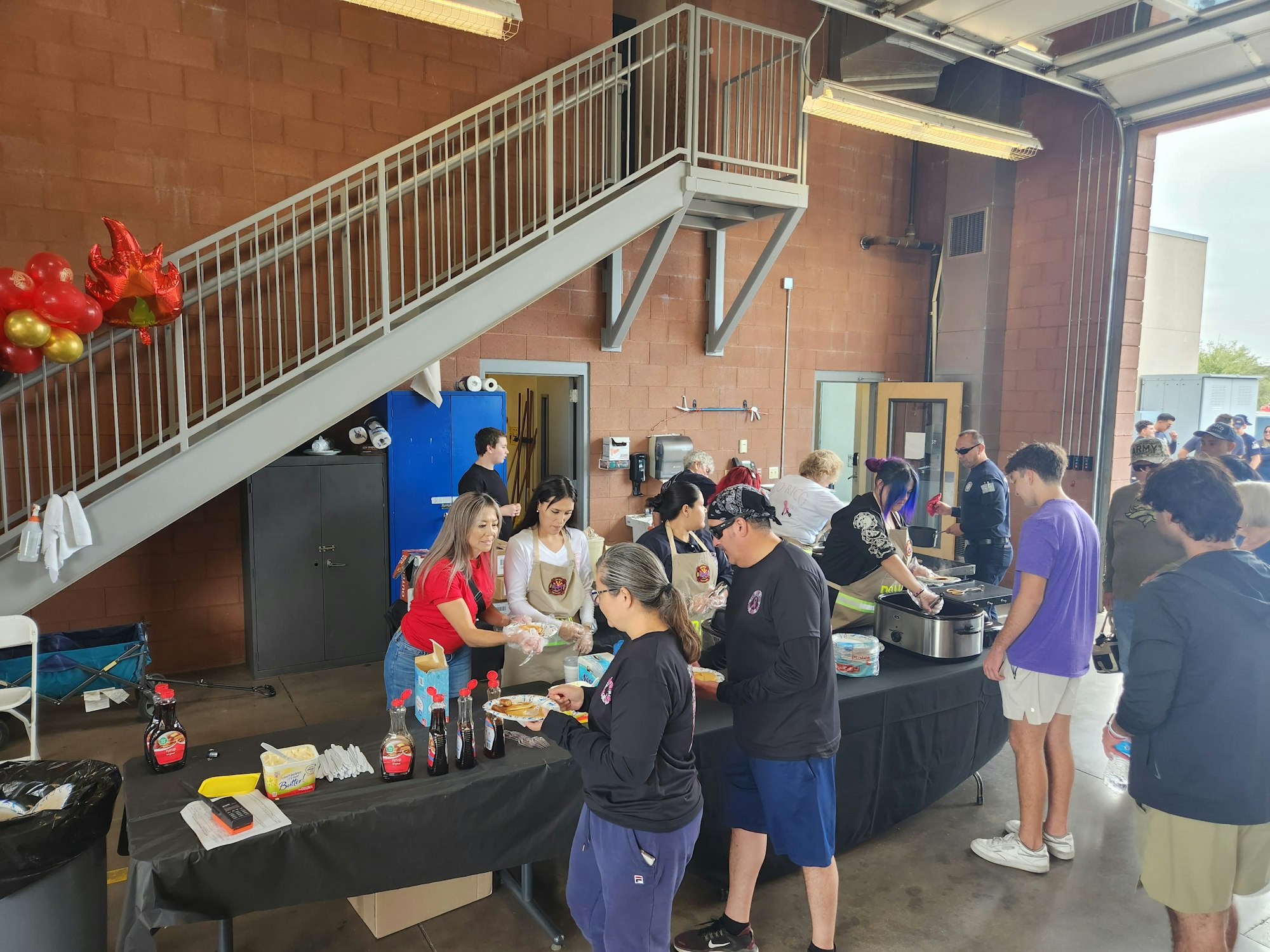 People gathered around a table with food and beverages, possibly at a community event or fundraiser in a garage-like space.