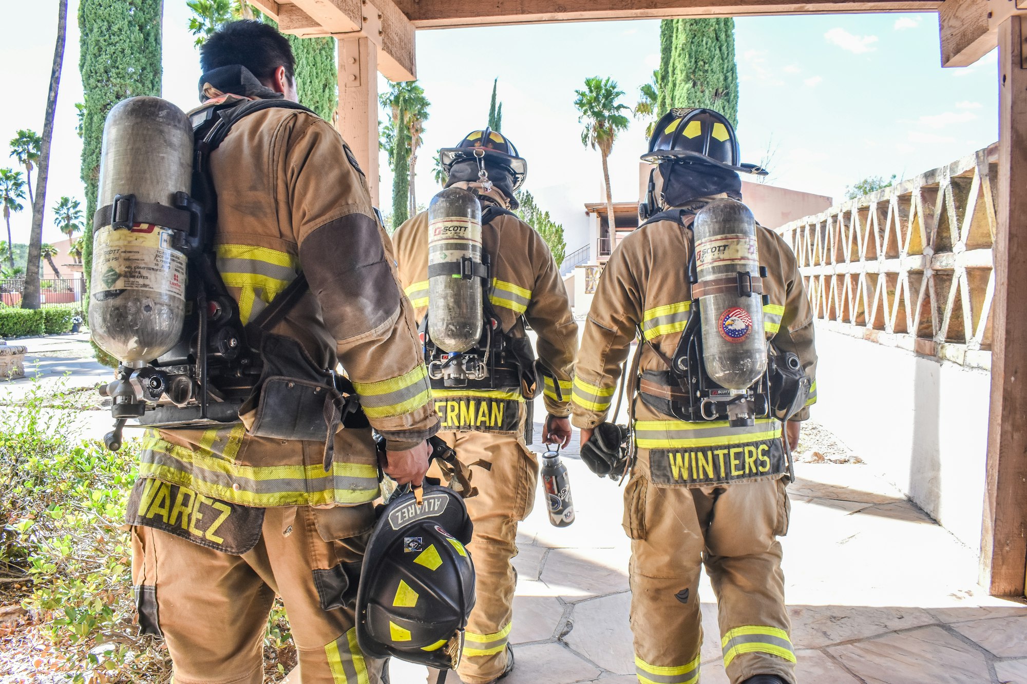 Three firefighters in gear with air tanks on their backs walking together.