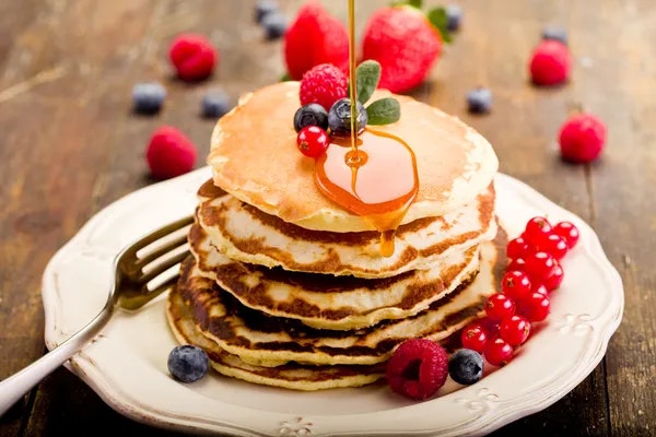 Stack of pancakes with syrup, garnished with berries (strawberries, raspberries, blueberries) on a white plate with a fork.