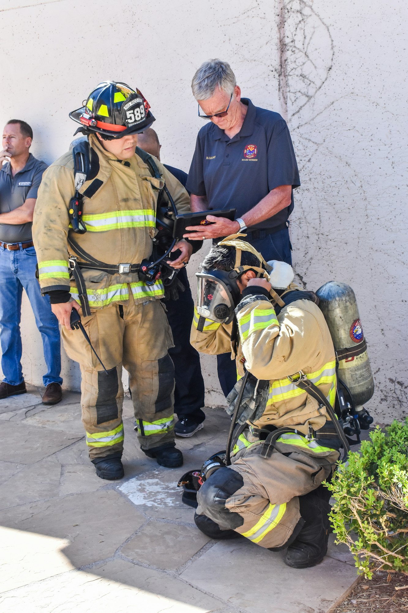 Two firefighters in gear with one kneeling and one standing next to a person holding a tablet.