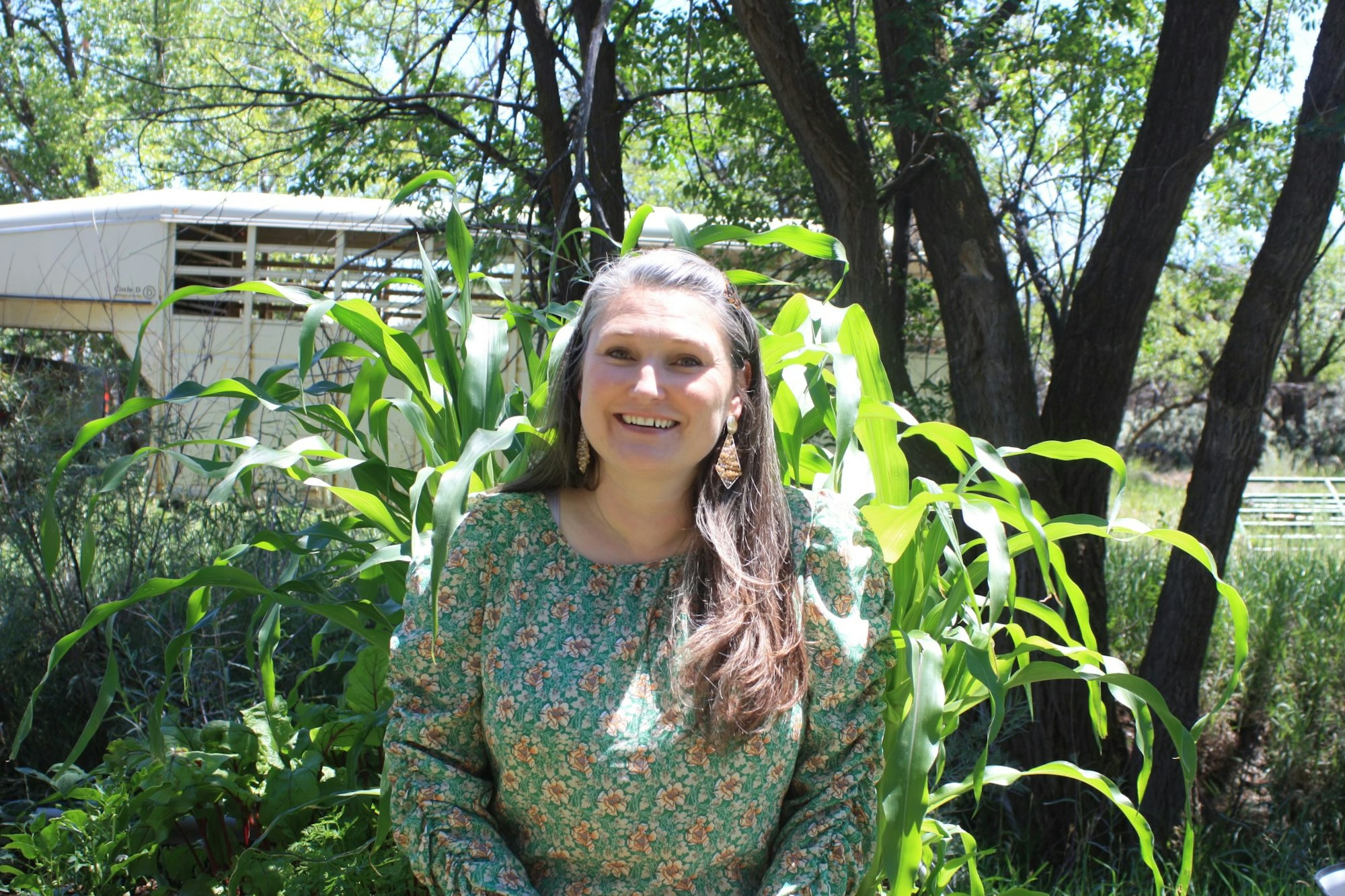 A smiling woman standing amidst greenery with trees and corn plants.