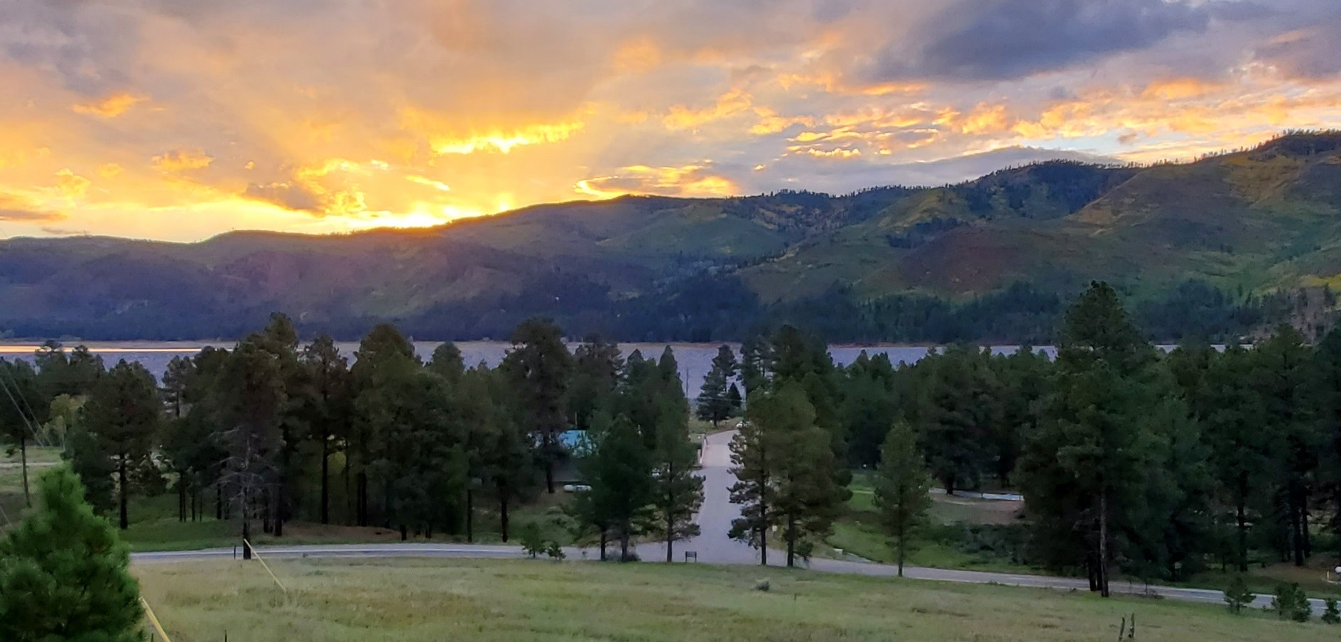 A scenic view of mountains at sunset, with trees in the foreground and a body of water in the distance. Beautiful colors in the sky.