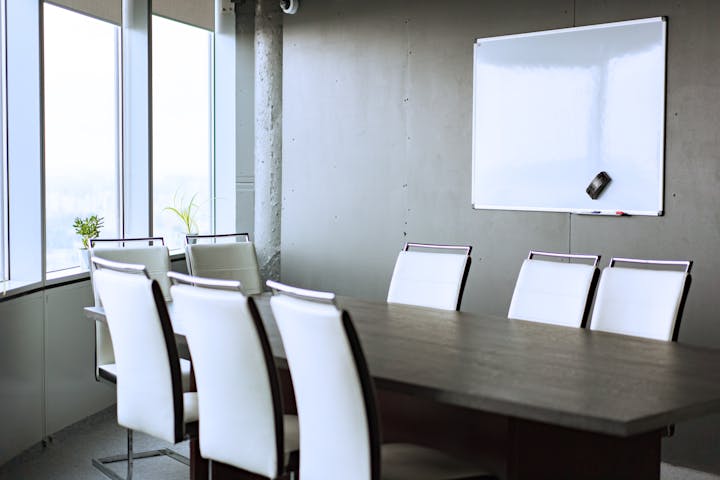 A bright conference room with a large table, white chairs, large windows, and a whiteboard on the wall.
