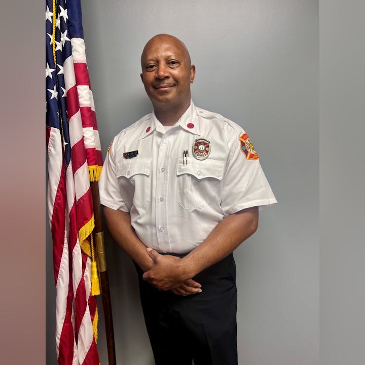 A uniformed firefighter stands beside a U.S. flag, smiling, in front of a gray background.