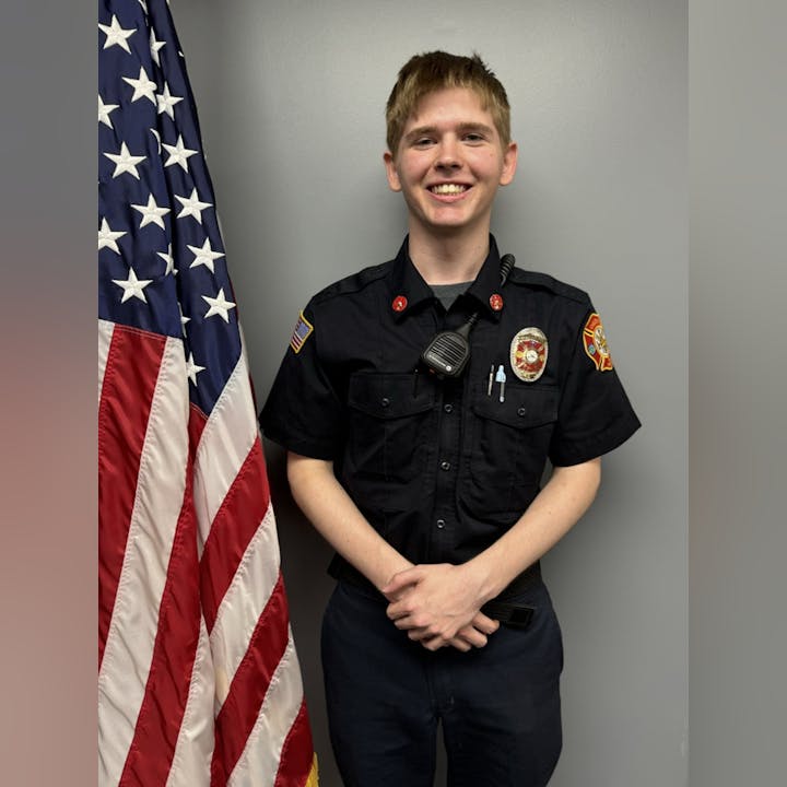 A person in a black uniform with badges stands beside a U.S. flag, smiling.