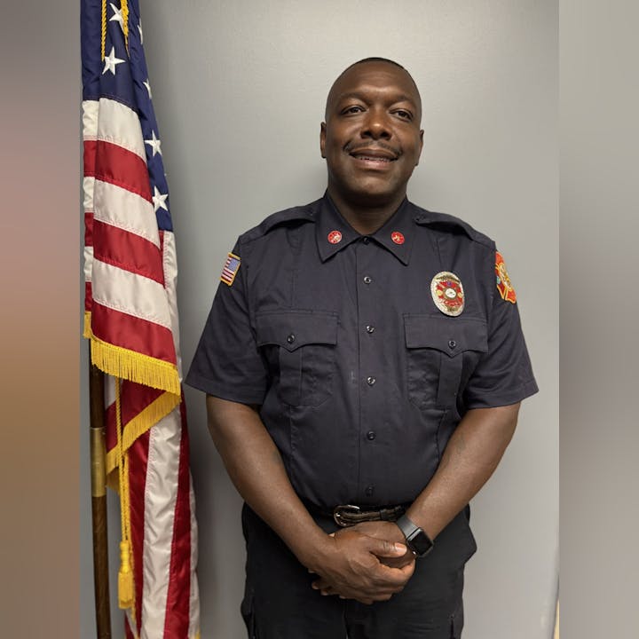 A person in a fire department uniform stands next to an American flag.