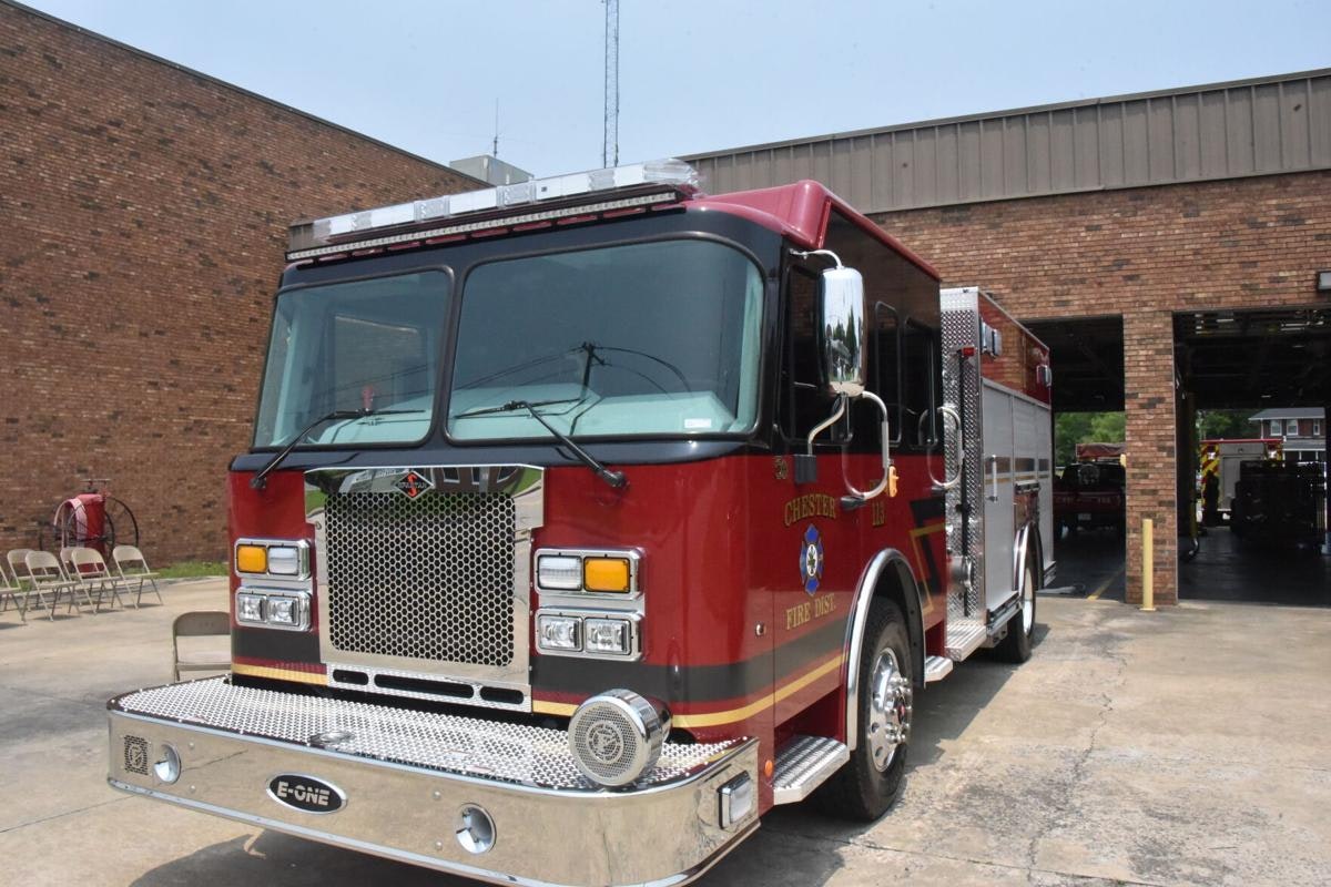 The image shows a red fire truck parked in front of a fire station, featuring a shiny front grille and emergency lights.