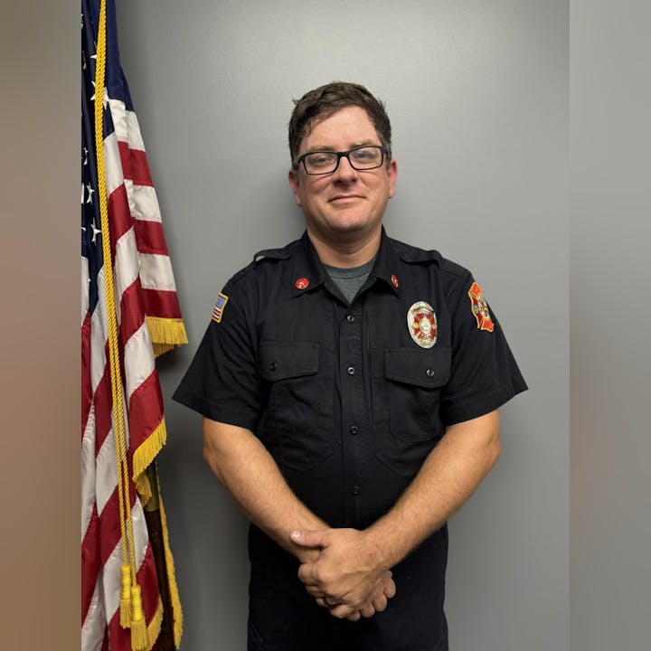A person in a firefighter uniform stands next to an American flag, against a gray background.