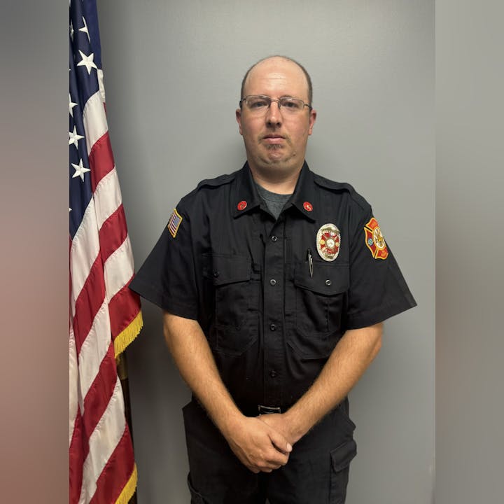 A person in a black uniform with fire department badges stands next to an American flag.